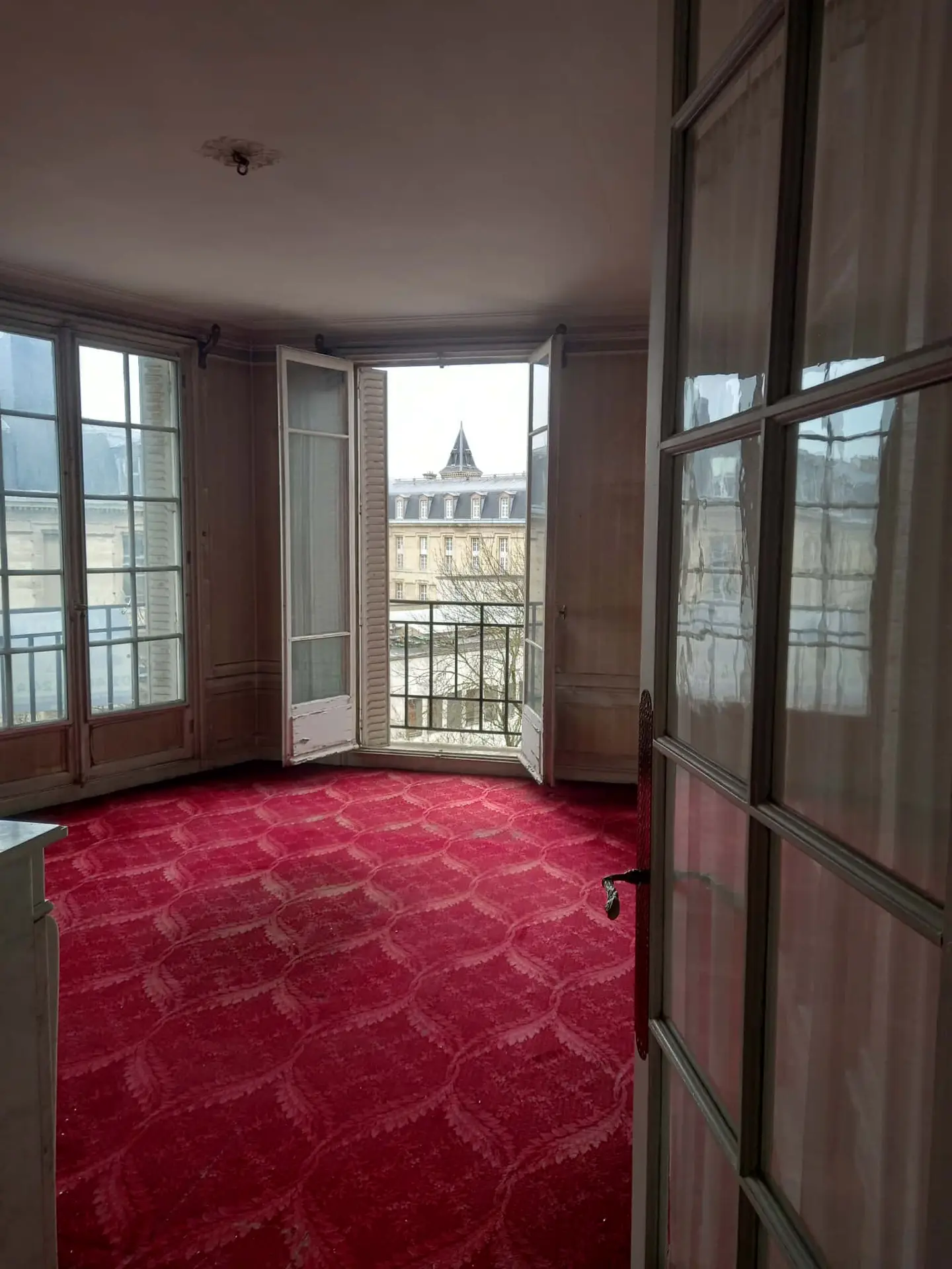Une pièce vide et lumineuse dans un appartement après débarras à Paris, avec une moquette rouge, de hautes fenêtres et des portes-fenêtres ouvertes menant à un petit balcon donnant sur les toits parisiens classiques, tandis que la lumière du soleil illumine doucement l'espace.