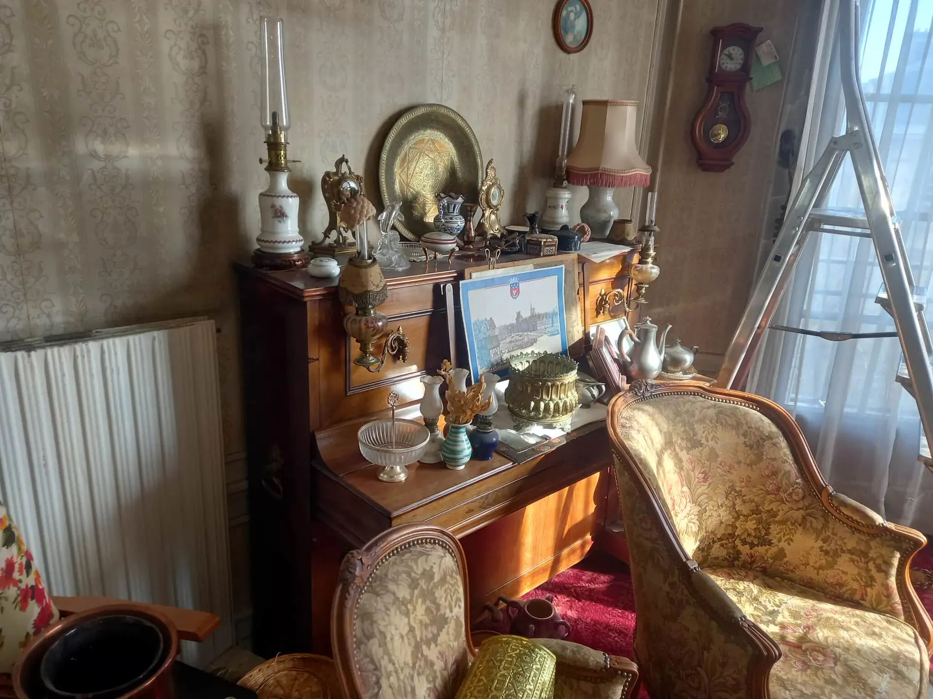 Une pièce encombrée avant débarras intégral, avec la lumière du soleil qui pénètre à l'intérieur. Un bureau en bois orné contient des vases, des théières, des chandeliers et des œuvres d'art encadrées. Des fauteuils à motifs sont assis à proximité ; une horloge murale et un radiateur ornent les murs tapissés. Paris et ile de france.