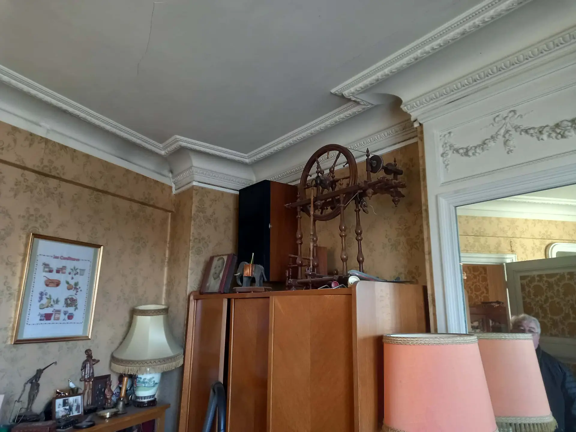 Une chambre vintage avec des moulures ornées, du papier peint floral et des meubles en bois dans un appartement à débarrasser à Paris et en Ile de France. Un rouet ancien orne une armoire ; des lampes, des tableaux encadrés, des photos et un miroir reflètent la tête d'une personne âgée.