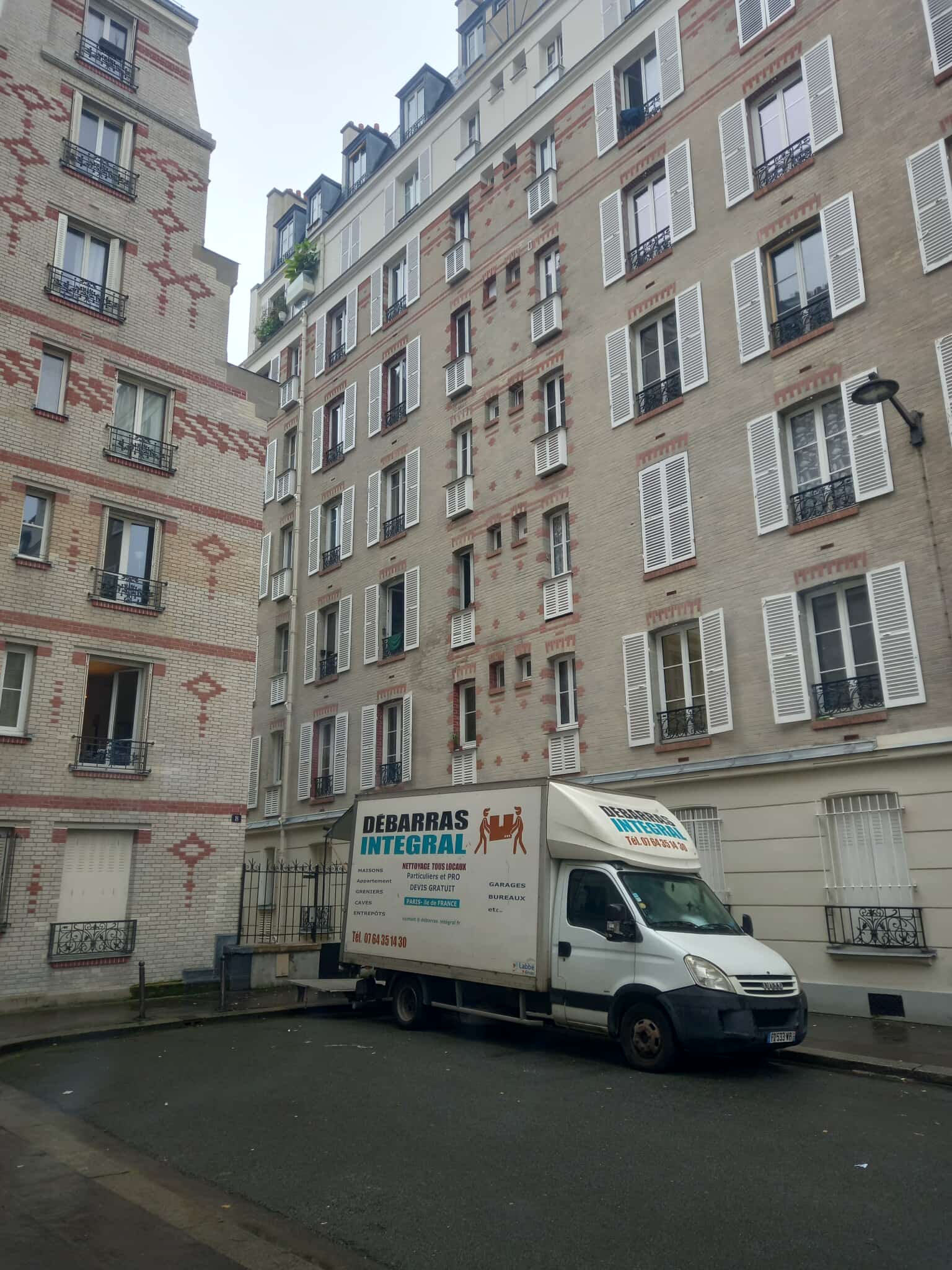 Un camion de débarras intégral à Paris, de couleur blanche, est garé dans une petite cour, entouré de grands immeubles parisiens en briques claires aux volets blancs. Le ciel gris et la rue vide ajoutent à l'atmosphère tranquille de la ville.