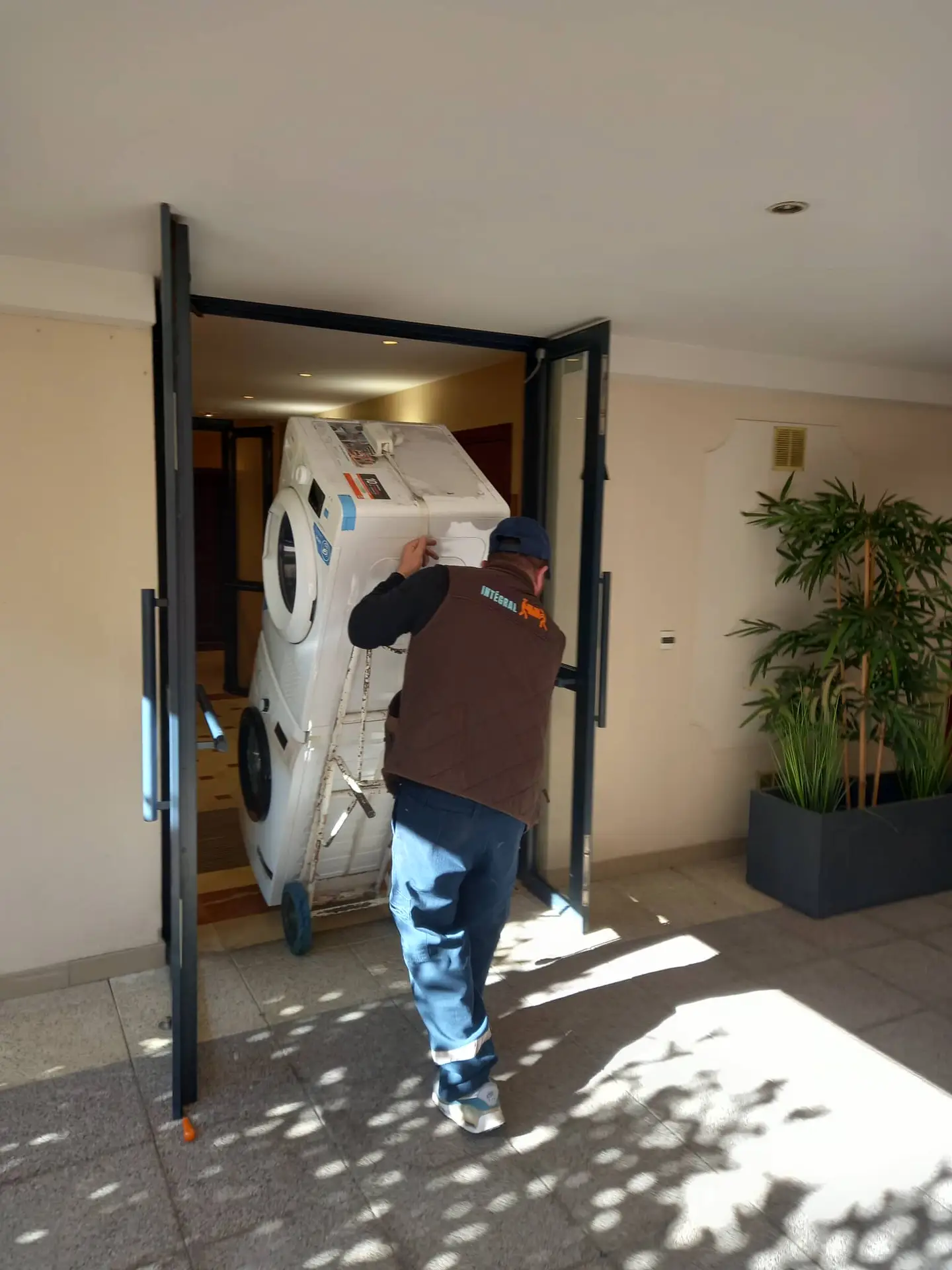 Un déménageur portant un gilet marron et une casquette bleue transporte une machine à laver inclinée sur un chariot à travers une porte étroite, la lumière du soleil projetant des ombres sur le sol. Parfait pour un débarras intégral ou pour débarrasser un appartement à Paris et en Île-de-France.