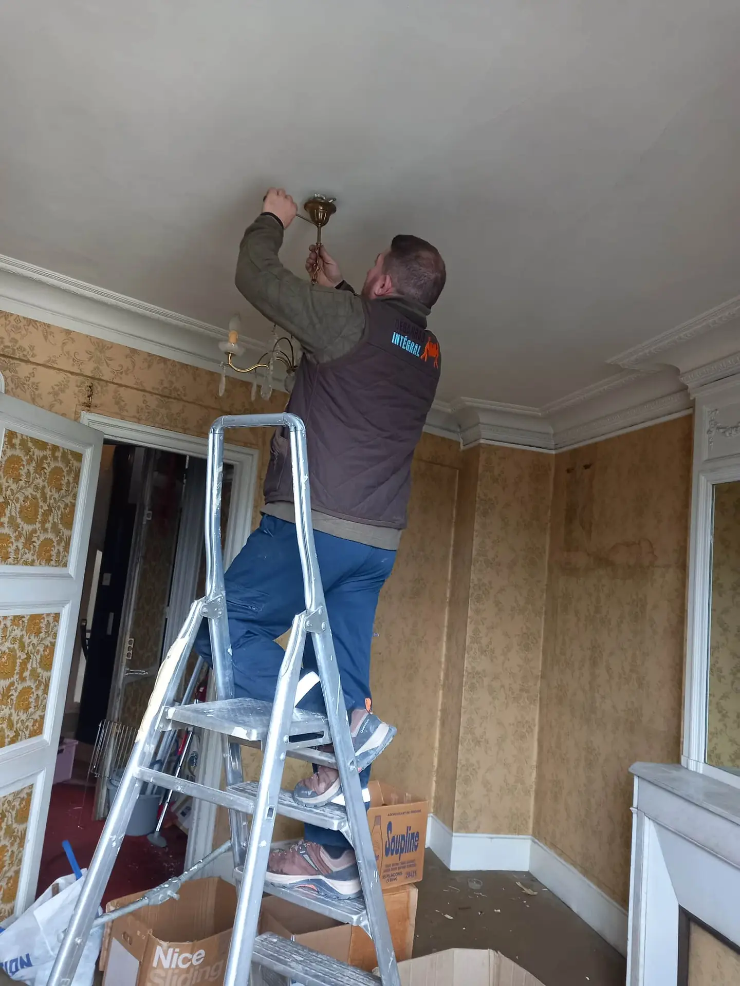 Un membre de débarras intégral se tient debout sur un escabeau, s'étirant pour installer un luminaire au plafond dans une pièce ancienne. Des boîtes et des outils d'une équipe de débarras intégral sont éparpillés sur le sol, prêts à débarrasser un appartement ou une maison à Paris et en Ile de France.
